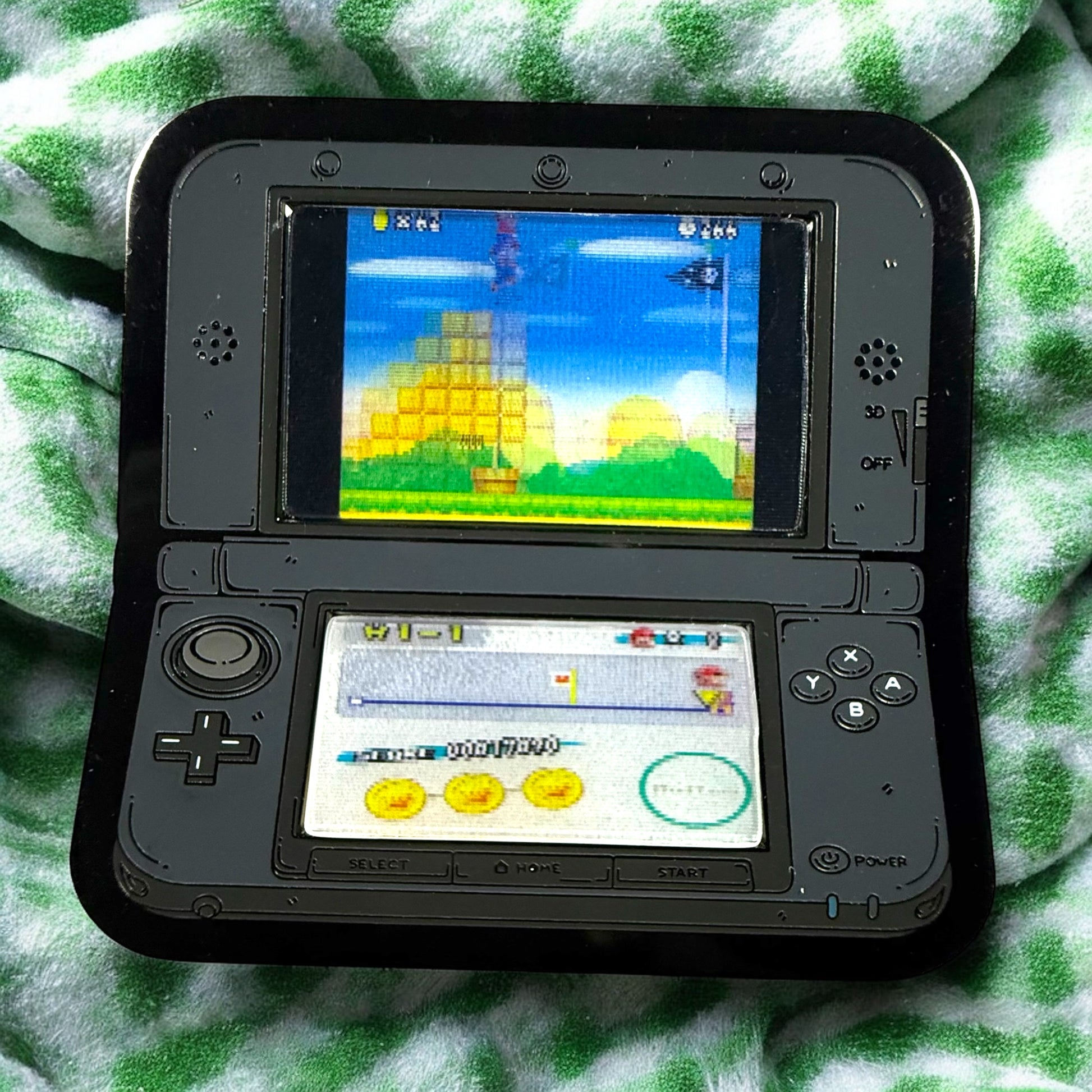 Nintendo DS gaming console with a game displayed on the screen, lying on a green and white checkered fabric.