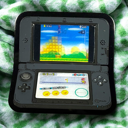 Nintendo DS gaming console with a game displayed on the screen, lying on a green and white checkered fabric.