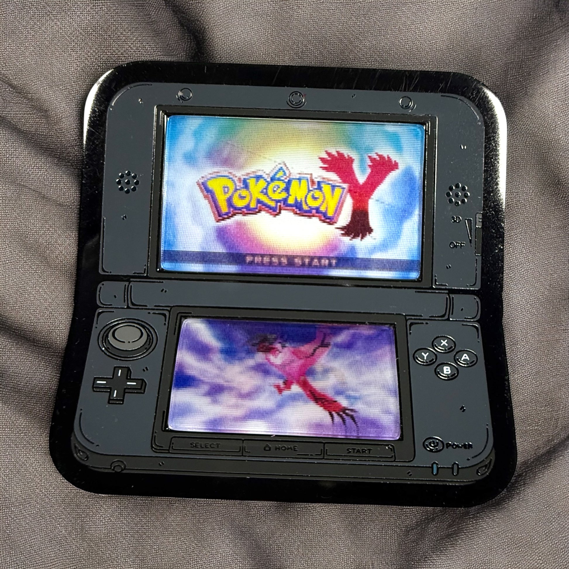 Nintendo 3DS gaming console displaying the 'Pokémon Y' title screen on a gray surface.