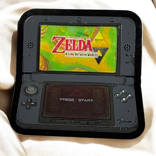 Black handheld gaming console displaying 'The Legend of Zelda: A Link Between Worlds' on a white surface.