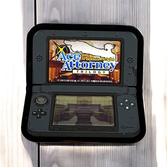 Black handheld gaming console displaying 'Ace Attorney Phoenix Wright Trilogy' on a wooden surface.