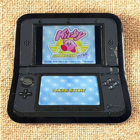 Black handheld gaming console displaying 'Kirby Super Star Ultra' on a textured beige surface